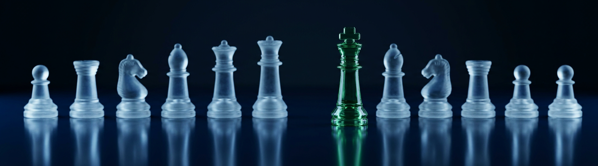 A row of clear glass chess pieces with a single green glass king stepping forward from the line